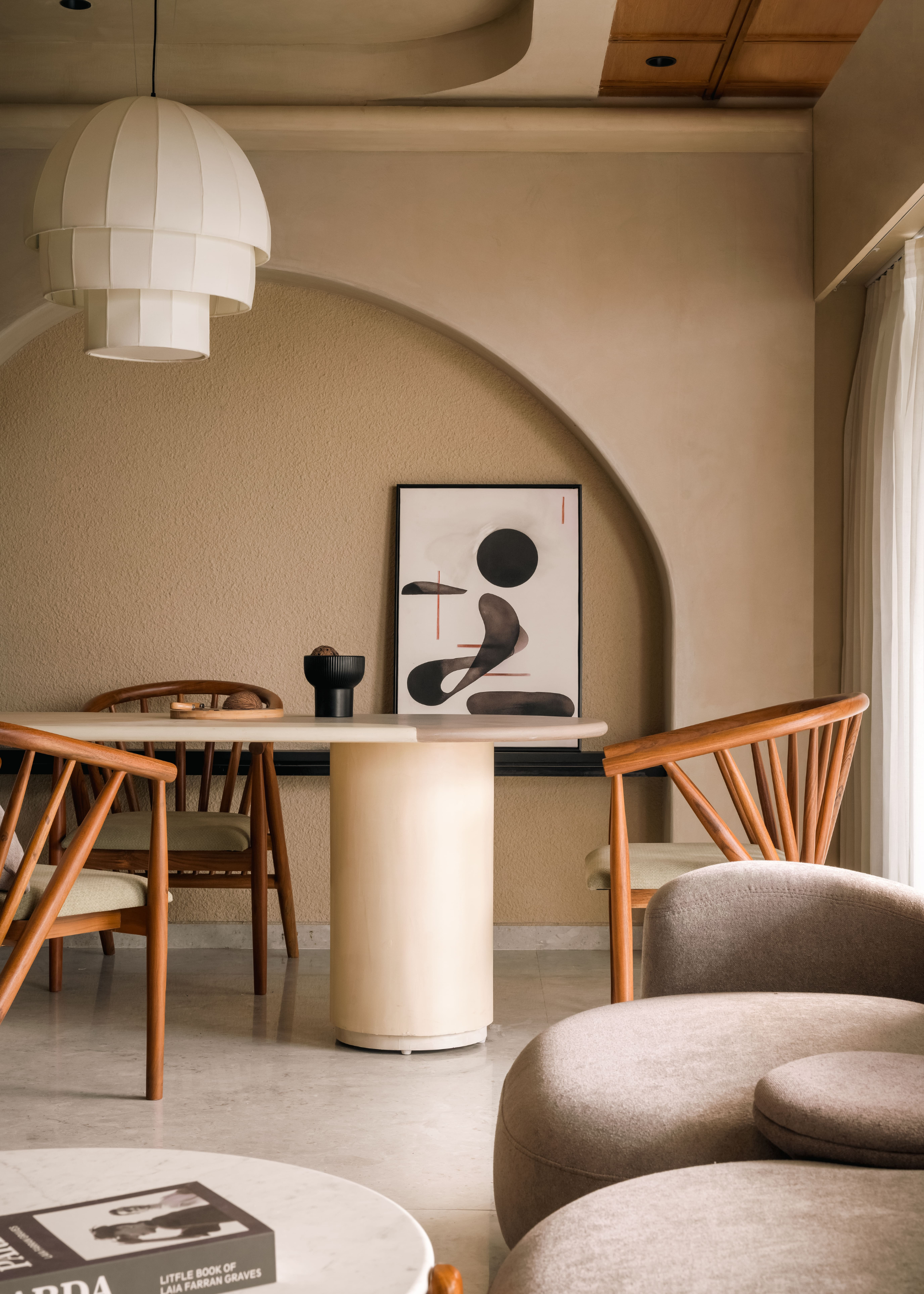 A living and dining room with beige walls, a leaning painting, a round dining table, wood armchairs, a paper lamp and a round coffee table by a sofa
