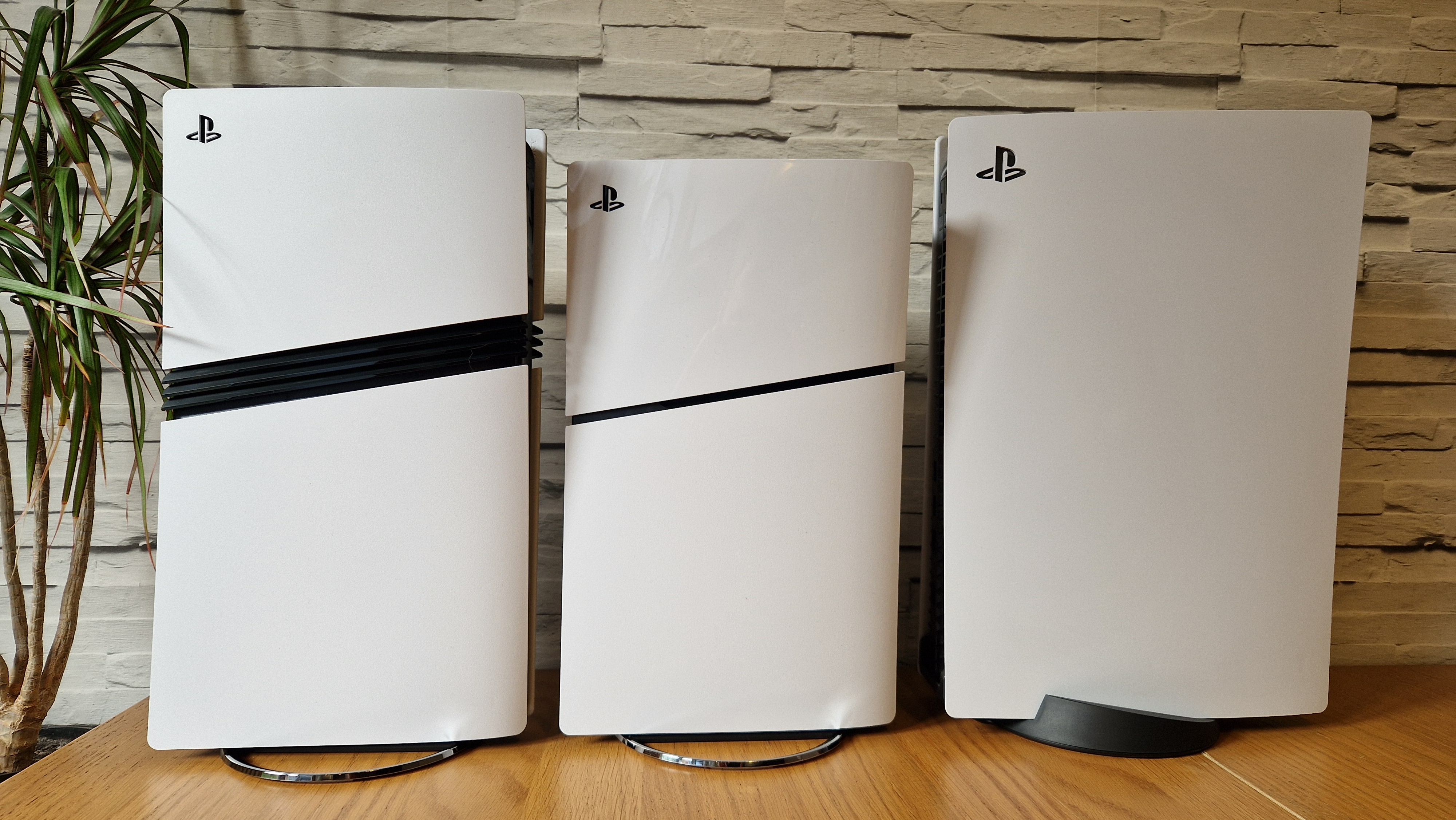 The PS5 Pro console next to a PS5 Slim and launch PS5 on a wooden desk in front of a white brick background and next to a potted plant