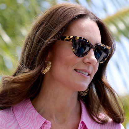 Kate Middleton wearing sunglasses and a pink collared dress with gold earrings looking to the right with palm trees behind her