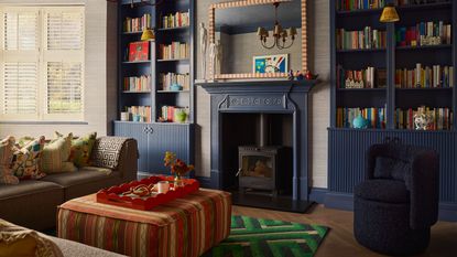 A cozy and colorful living room with dark blue built-in bookshelves and trim, filled with colorful books. There is a beige L-shaped sofa with an orange-patterned ottoman in the middle and a green geometric print rug underneath.