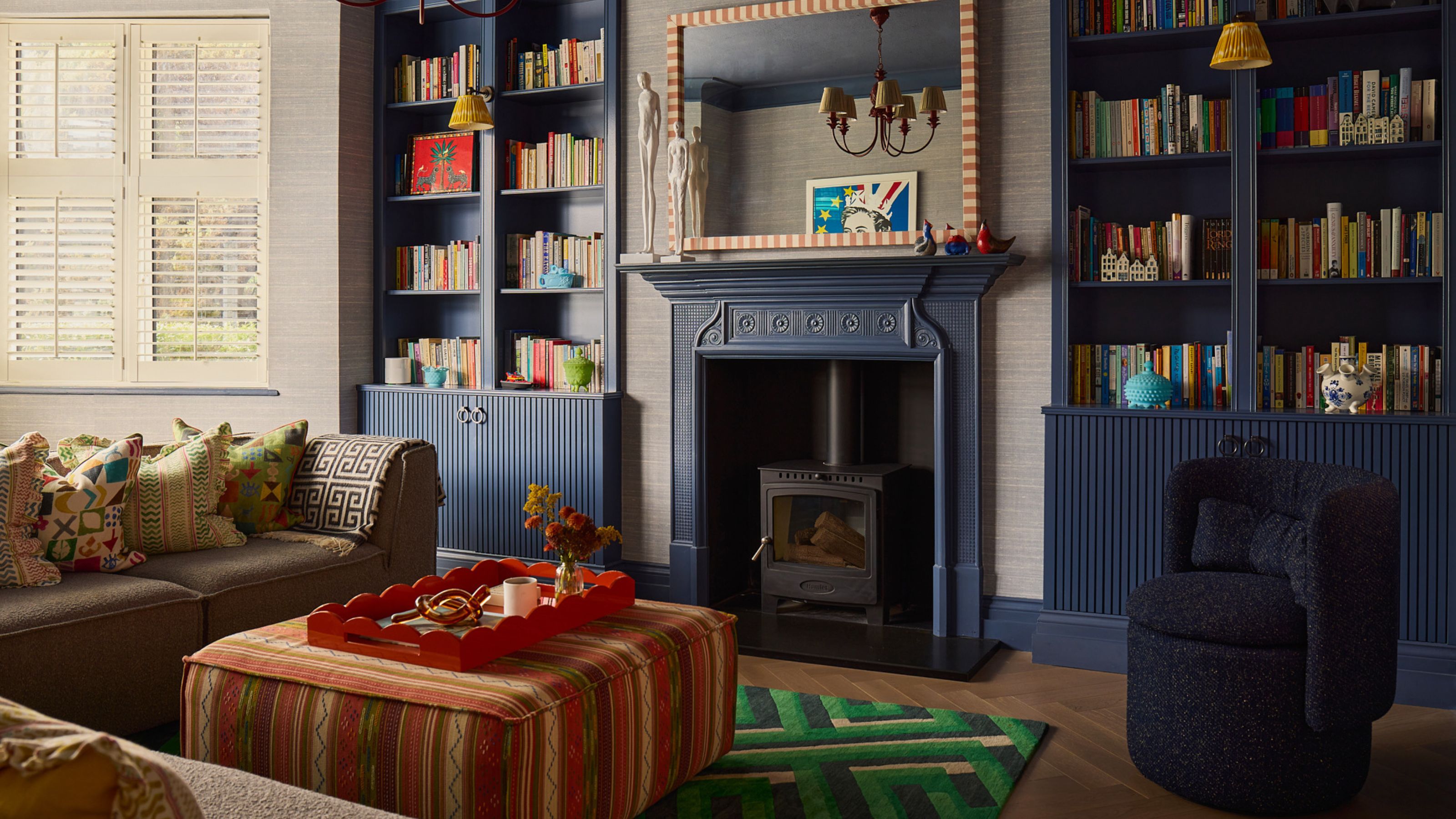 A cozy and colorful living room with dark blue built-in bookshelves and trim, filled with colorful books. There is a beige L-shaped sofa with an orange-patterned ottoman in the middle and a green geometric print rug underneath.