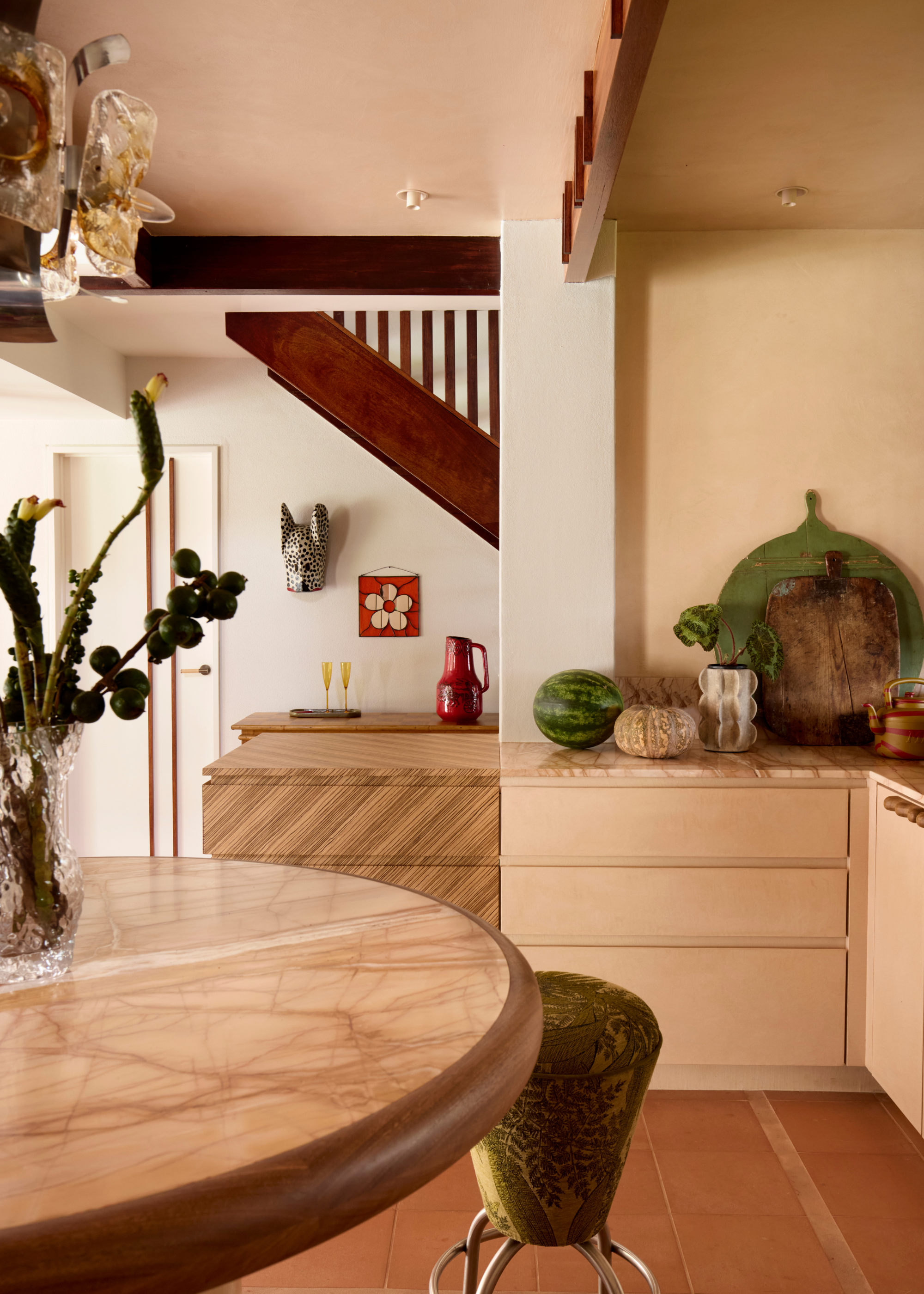 The connecting space between an L-shape kitchen counter and circular kitchen island with a green cone bar stool with chrome legs, abstract cacti-like foliage in a vase on the island, and timber and veneer joinery throughout along with terracotta flooring