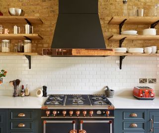 A close up of a large range cooker with brass knobs and a large extractor fan. The wall is small white brick tiles and exposed brick above