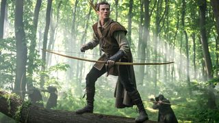 Robin Hood stands on a tree trunk with a bow and arrow with two hooded archers behind him