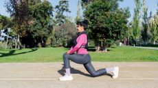 Woman doing walking lunges along sunny path in park wearing workout clothes