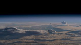 A mantle of clouds blankets the horizon looking down at a section of Earth from space. Tall cloud pillars shoot up from various spots.