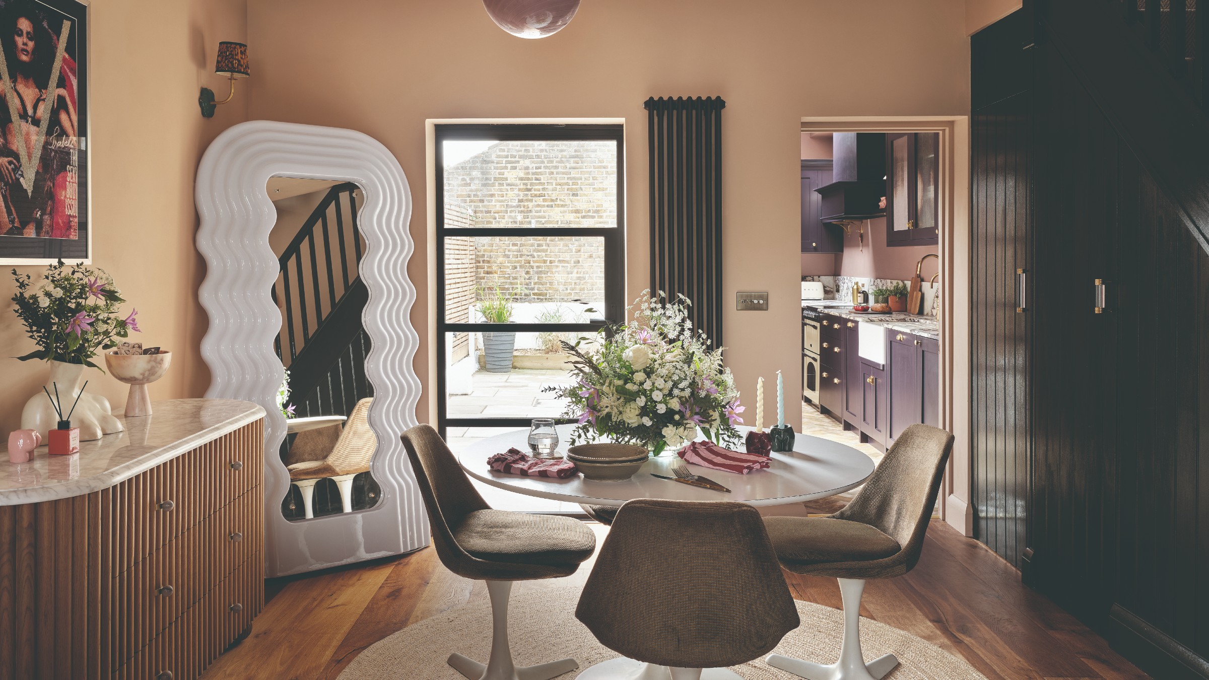 A pink-painted dining area with a white round dining table, a reede console table and a wavy full-length mirror in the corner