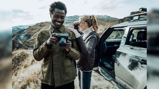 Two people outside in the hills with cameras and a car