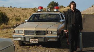 Javier Bardem as the hitman Anton Chigurh standing next to a tan sheriff car in the movie No Country for Old Men.