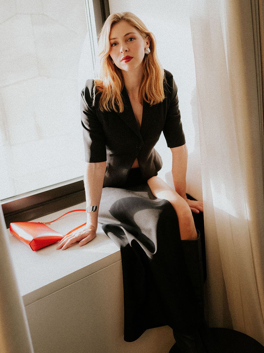 Actress Hermione Corfield, star of the series Outlander: Blood of My Blood. She is sitting in front of a window in the corner of a room wearing a black blazer and skirt with knee-high boots.