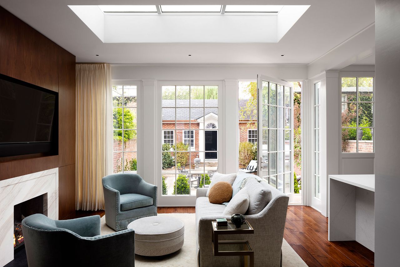 family room with blue club chairs and off-white sofa and round footstool with fire lit and wallmounted TV and pivot door open to backyard and big rooflantern above cream rug on dark wood floor