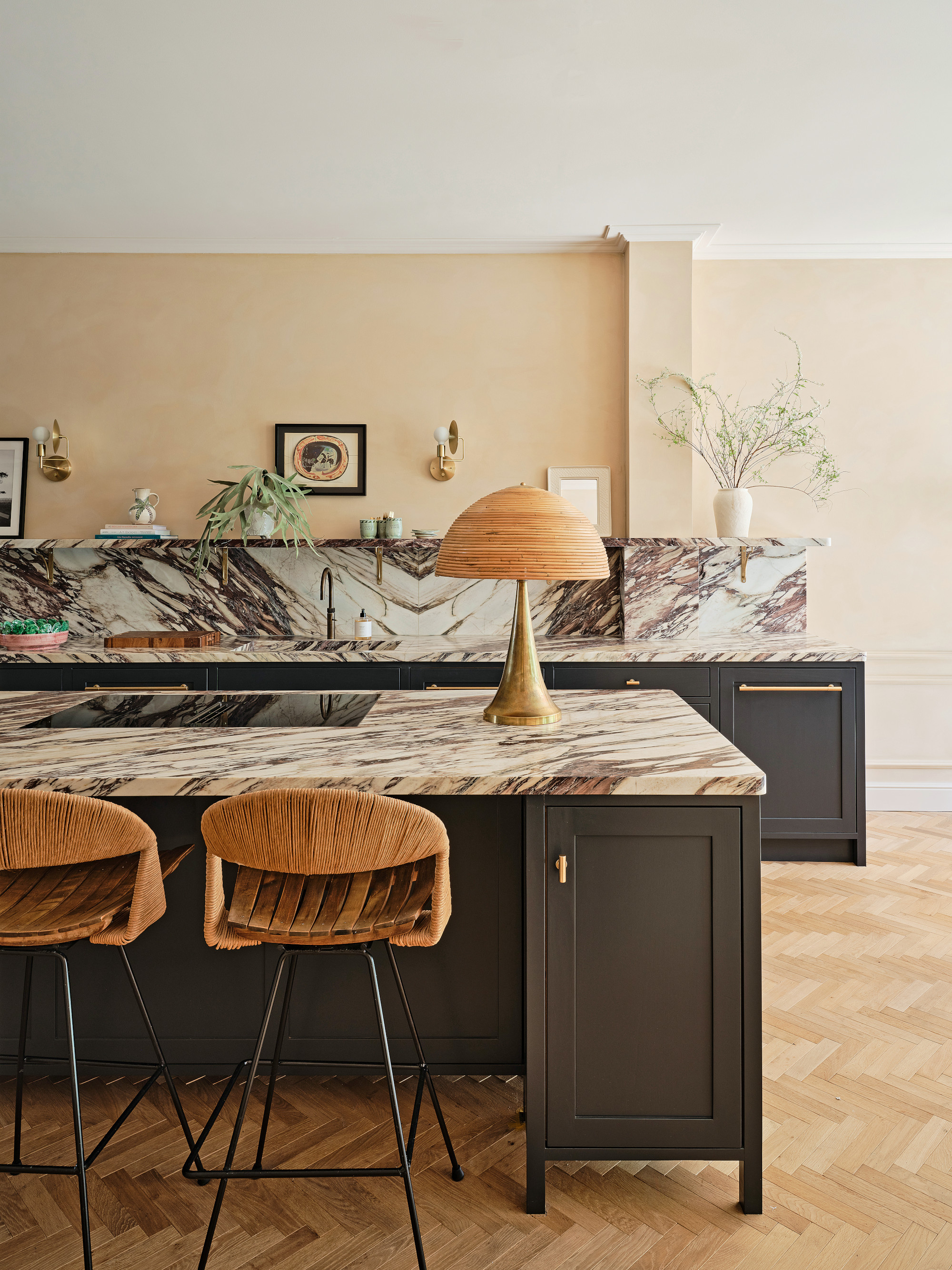 Black Shaker-style kitchen with burgundy and white marble countertop, woven bar stools and table lamp with brass base, cream walls and herringbone wood floor