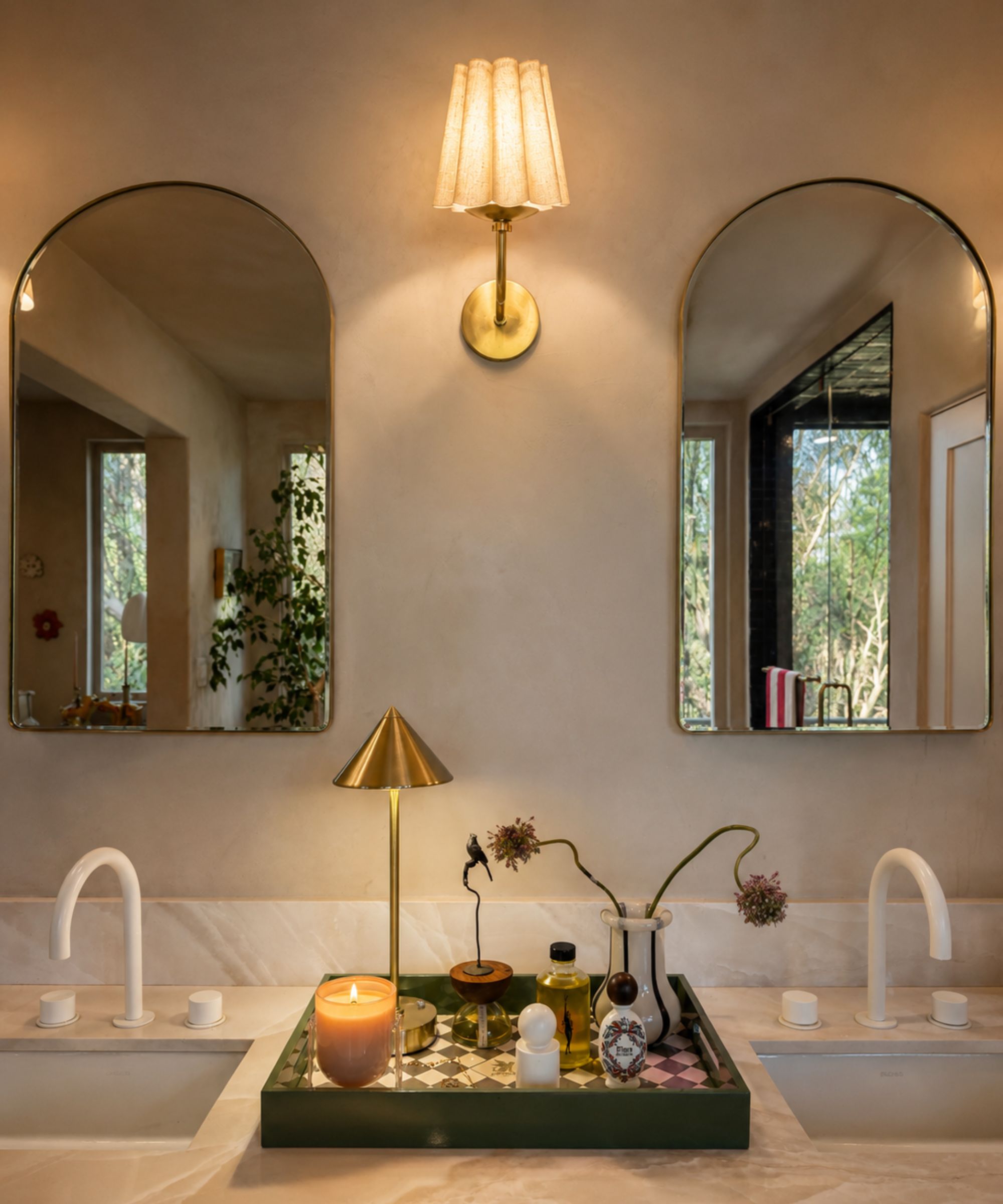 Modern bathroom with double-vanity, two arch mirrors, marble countertop and backsplash, large tray with lamp, vase of flowers, perfumers. Wall lamp inbetween mirrs