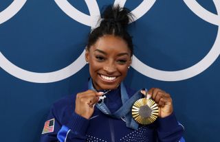 Simone Biles holds up a goat pendant necklace and a gold medal after winning the women's all around