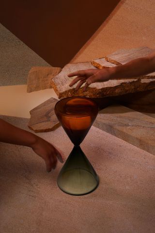 An hourglass in the tones of orange and green is placed against a sandy background as two hands get closer to it.