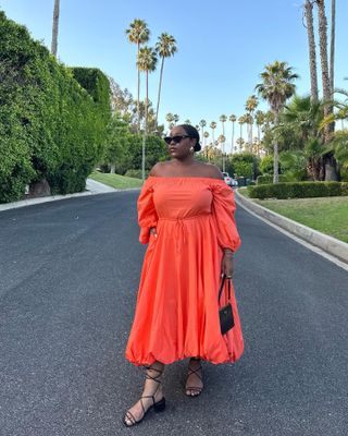 Aniyah wears a orange flowy sundress with sunglasses, black sandals, and a black handbag.