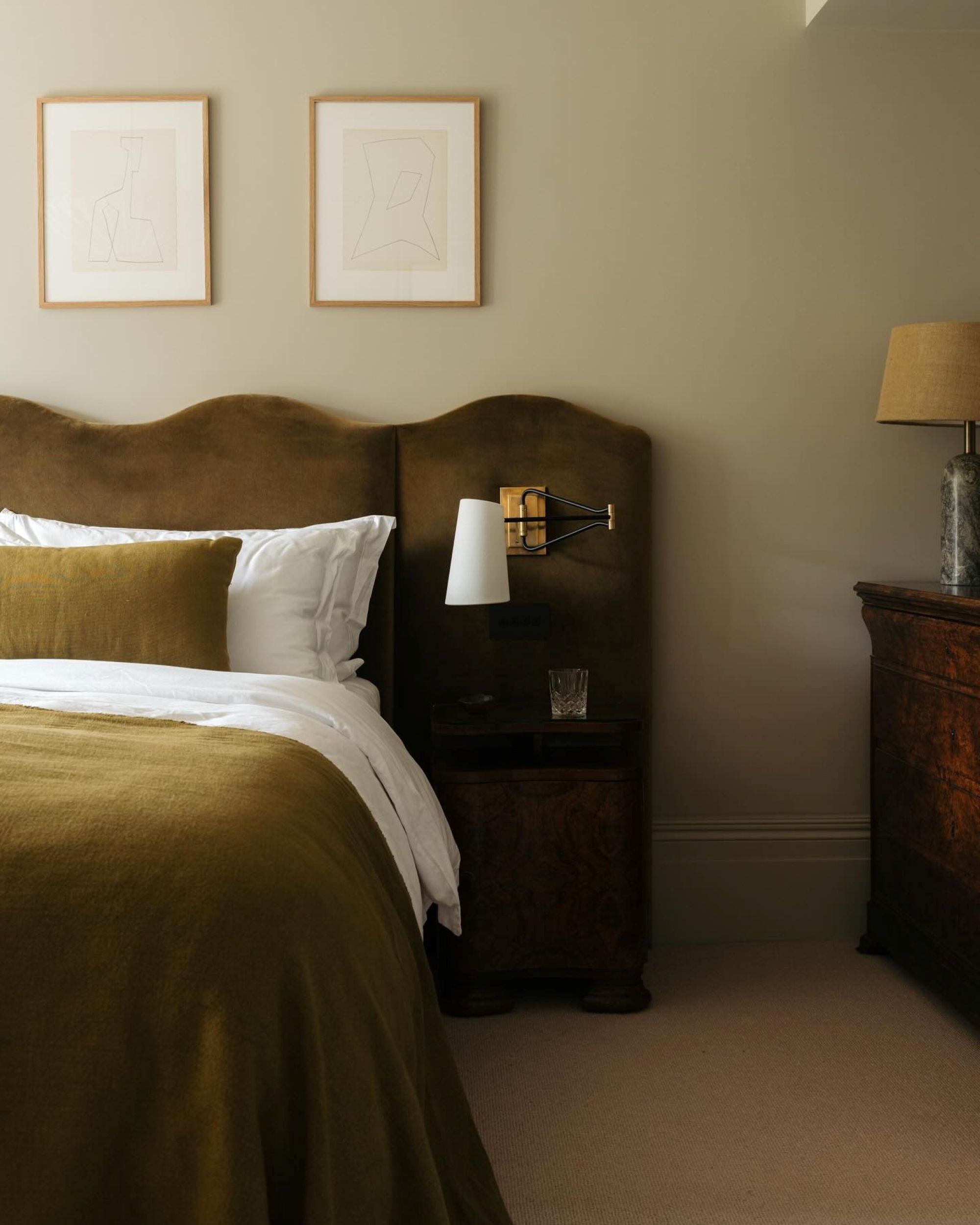a bedroom with a wavy velvet green headboard, a bed with white sheets and an olive green top sheet, and olive green pillow, and soft beige paint on the walls. Above the bed is two framed images