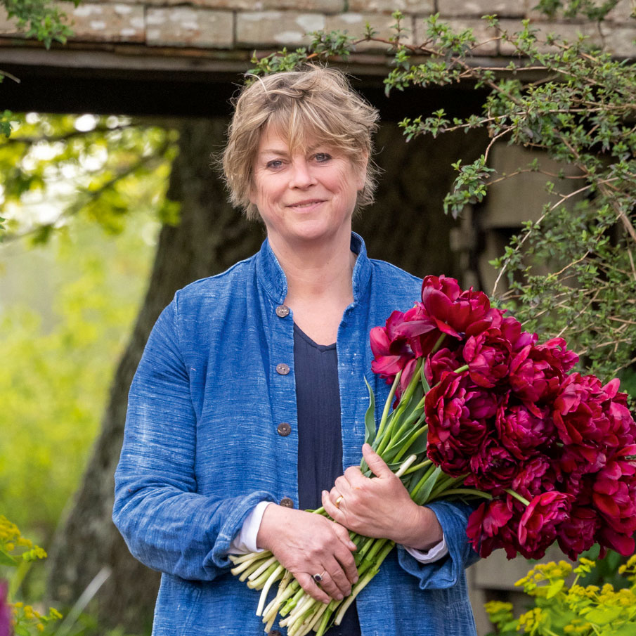 Sarah Raven gardener facing the camer holding a bunch of dark pink peonies