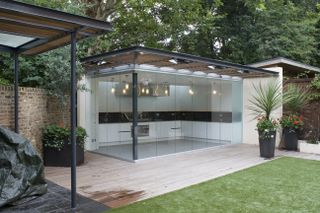 a totally glass garden room with wooden slat room, with an all-white kitchen inside with black counters and backsplash
