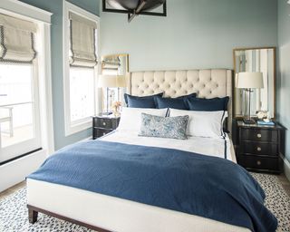 Blue bedroom with pale blue painted walls, cream upholstered headboard, white linen with blue throw and cushions, dark wood bedside tables, matching bedside lamps with white shades, two large mirrors either side of bed, animal print rug