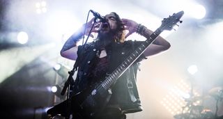 Robb Flynn of Machine Head cups his hands to his ears as he performs with a V-style electric guitar on a stage bathed in white light.