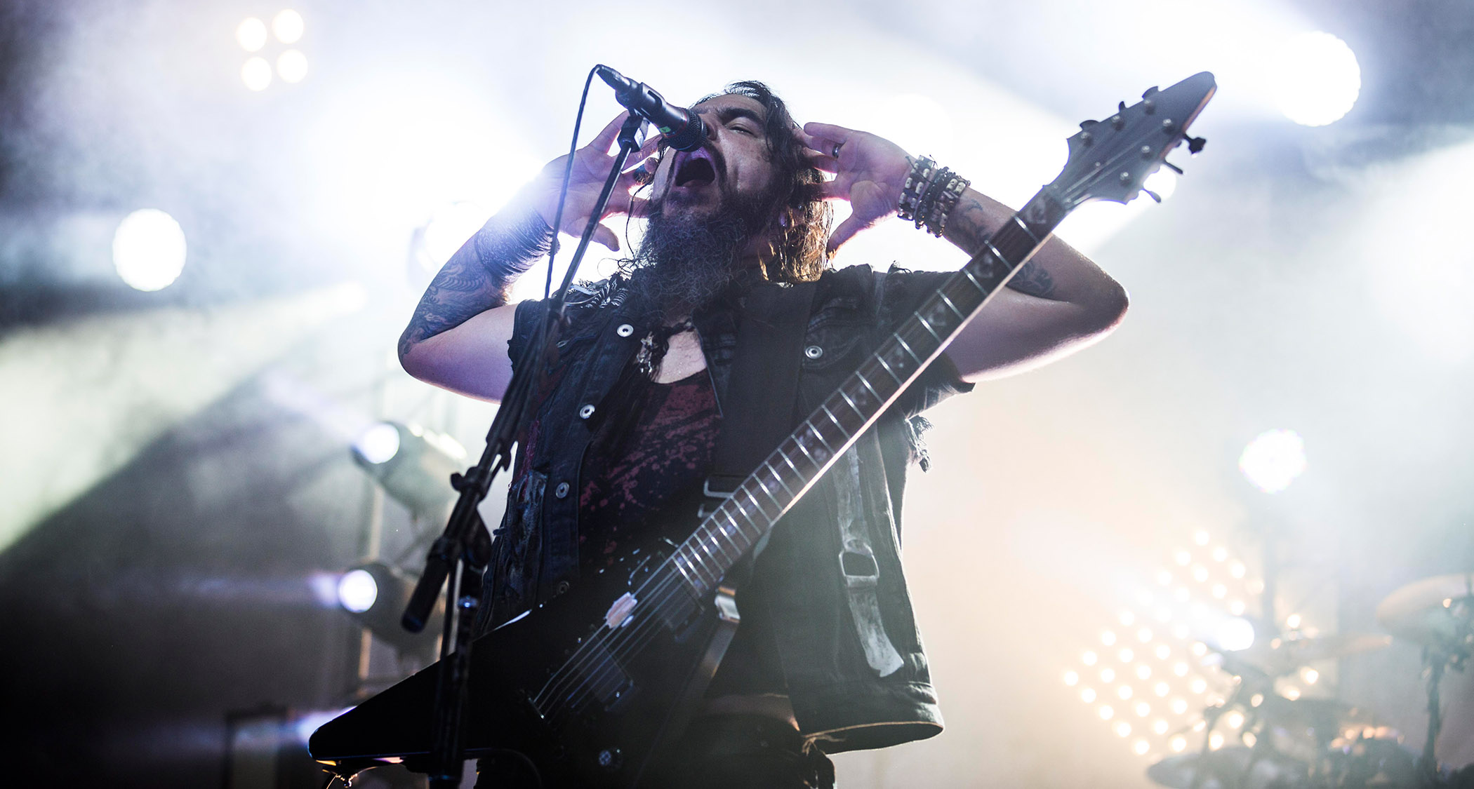 Robb Flynn of Machine Head cups his hands to his ears as he performs with a V-style electric guitar on a stage bathed in white light.