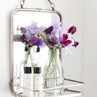 A hanging mirror on the wall with a shelf holding a vase of fresh flowers and skincare products