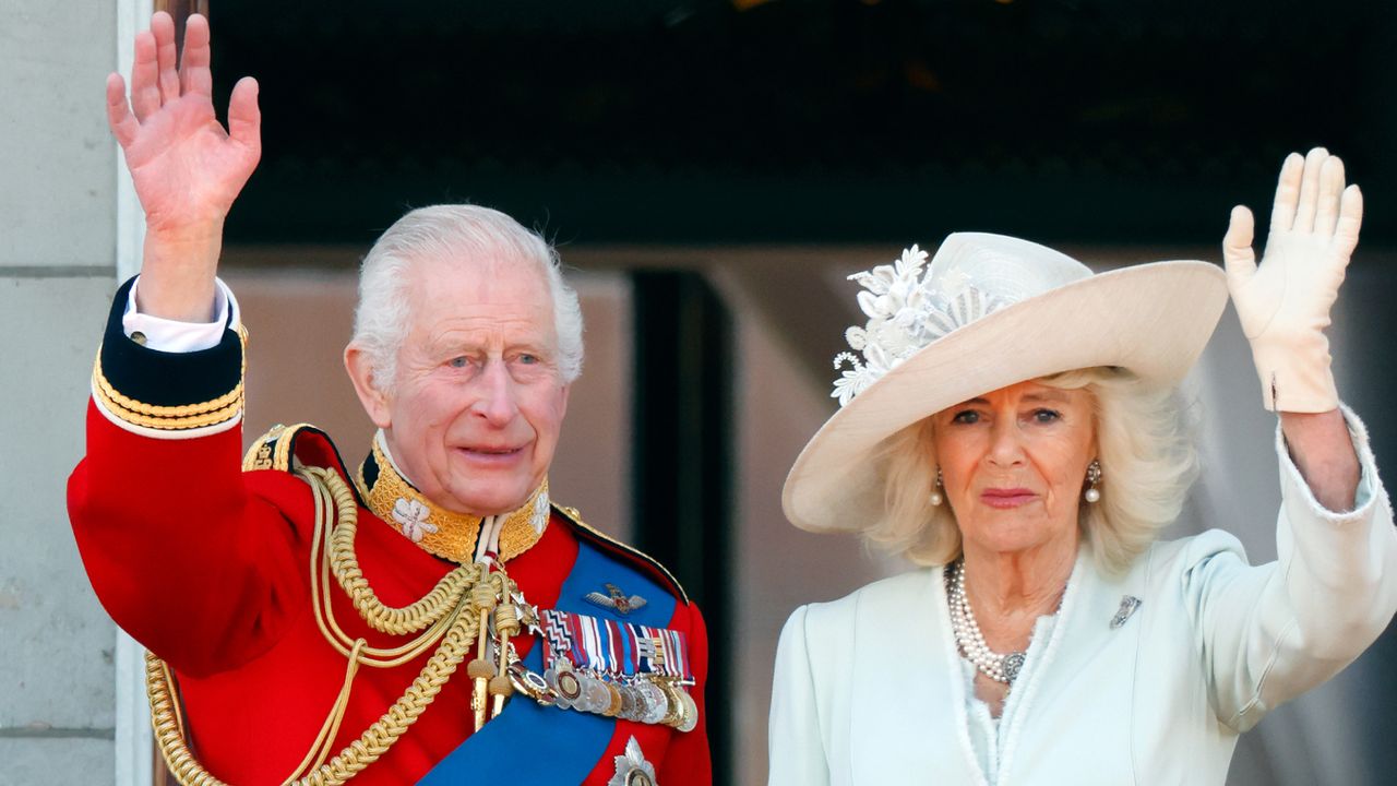 King Charles wearing a red military uniform and Queen Camilla wearing a mint green coat and matching hat waving 