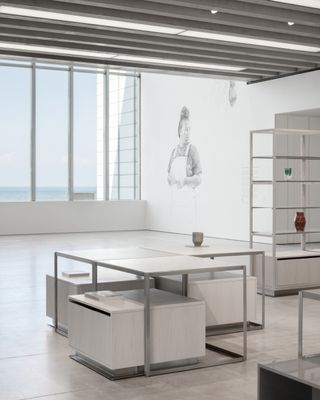 Alternative interior view at the Turner Contemporary shop featuring white walls, light grey flooring, white and grey shelving units and tables with single drawer units underneath, rectangular ceiling lights and windows offering a view of the sky and sea. There is an illustration of a person on the wall by the windows