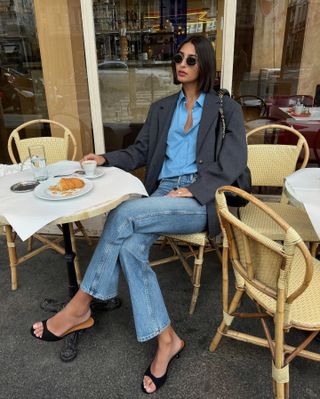 French fashion influencer Salome Mory sitting at an outdoor Paris cafe wearing round sunglasses, a gray blazer, blue button-down shirt, straight-leg jeans, and open-toe mule sandals.