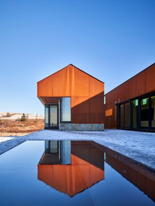 Exterior view of the Corten steel and stone pavilion structures at Smith House under a clear blue sky. There is a swimming pool outside surrounded by a snow covered area and there is brown grassy land and trees nearby