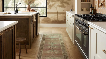Antique-style patterned rug lining the tiled floor of a charming kitchen area, which features a glossy black stove, rustic wooden stool, and neutral nature murals decorating the walls.