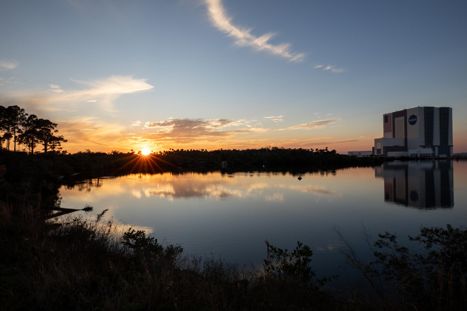 a still body of water rests, flanked on the left by overbrush and a few trees. The shoreline curves back toward a tall cube building, reflected in the water across from the rising sun on the horizon.