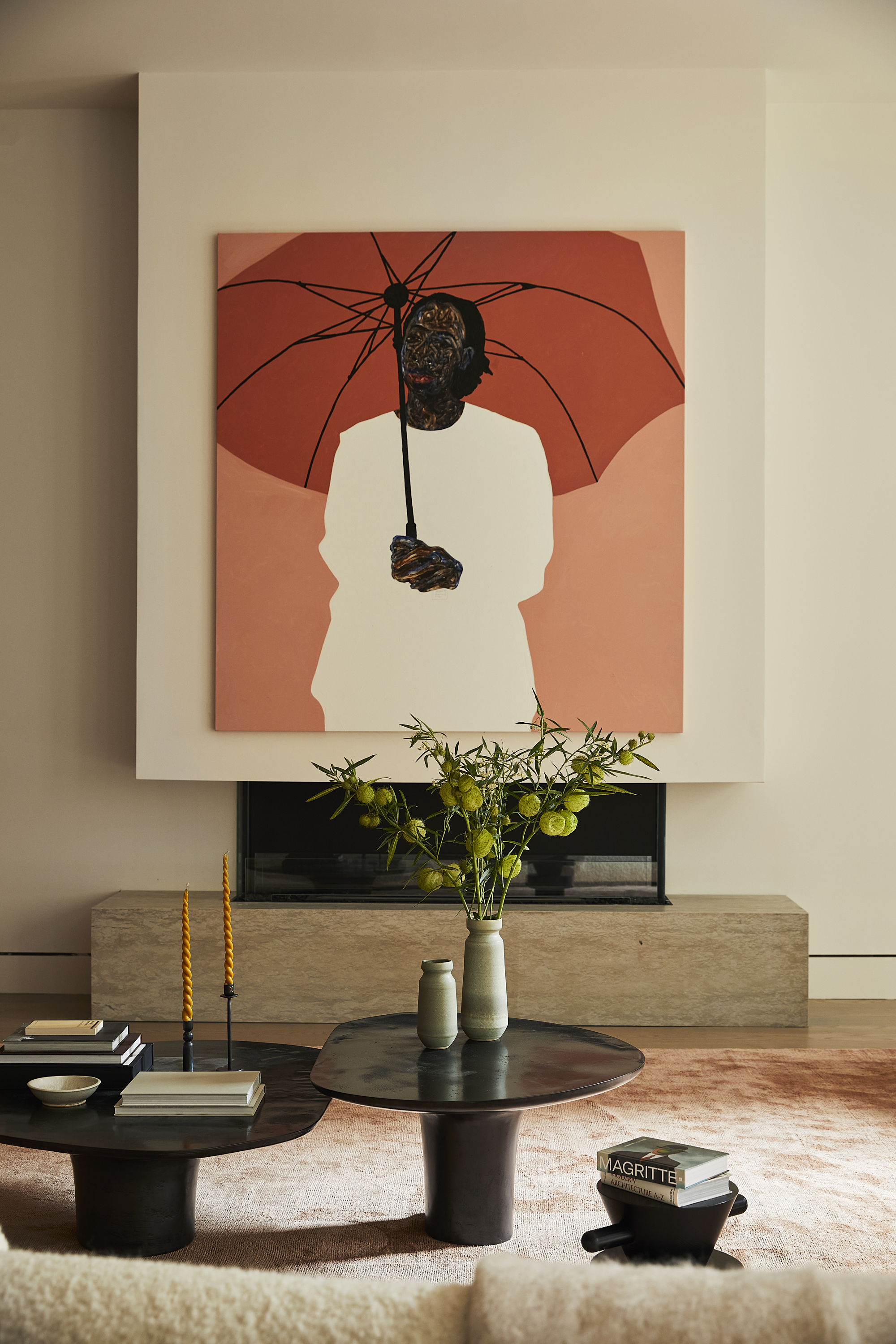 beige living room with large orange artwork hung above low-marble gas fireplace, with orange rug, two black nesting coffee tables, a stack of books, and a beige sofa