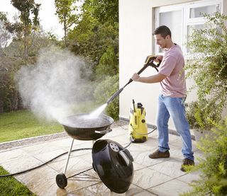 A picture of a male adult wearing blue denim jeans using a Karcher pressure washer to clean a barbecue outside in his backyard