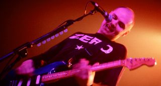 Billy Corgan of the Smashing Pumpkins performing with a Zero T-shirt and Fender Strat, on a stage illuminated in orange with pink lights on his face.