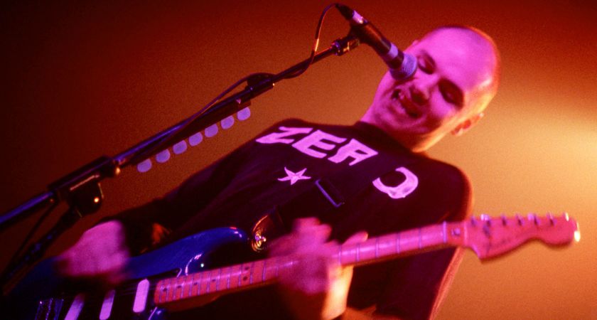 Billy Corgan of the Smashing Pumpkins performing with a Zero T-shirt and Fender Strat, on a stage illuminated in orange with pink lights on his face.