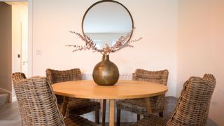 A round wood dining table with wicker chairs, gold coloured round vase with dried flowers and mirror in a dining room within a home