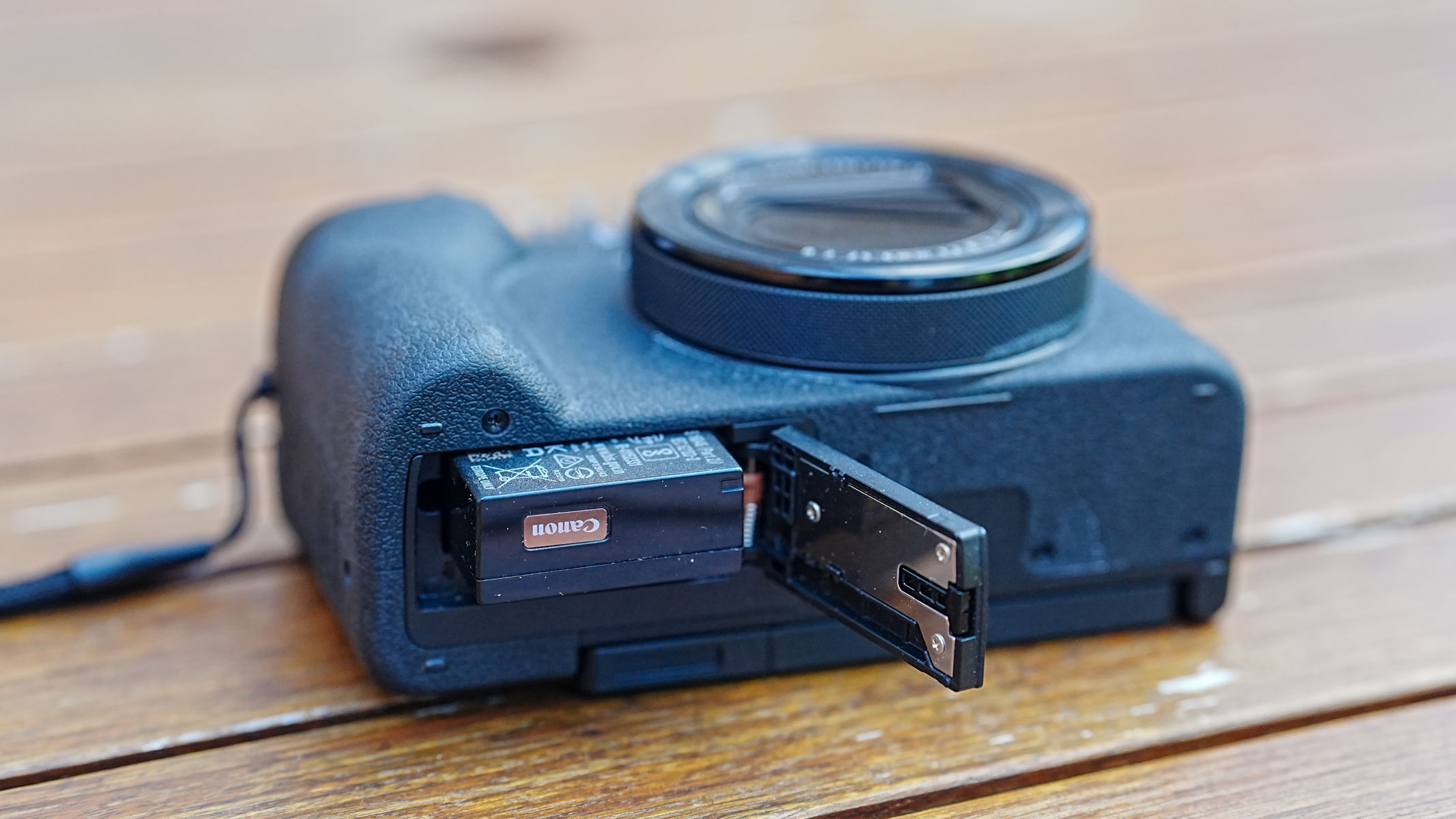 The Canon Powershot V1 on a wooden table with the battery compartment open with battery poking out.