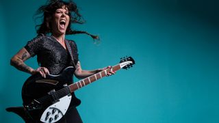 A photo of Lilly Hiatt jumping against a deep blue background while wearing her black Rickenbacker electric guitar