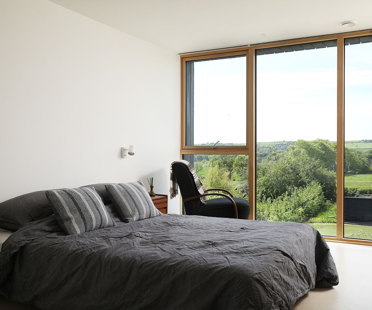 A double bed with crumpled cotton duvet cover in a dark grey colour. There is a wall of floor-to-ceiling-glazing looking out towards views of the countryside. A dark armchair sits next to the window angled outwards so the view might be enjoyed.