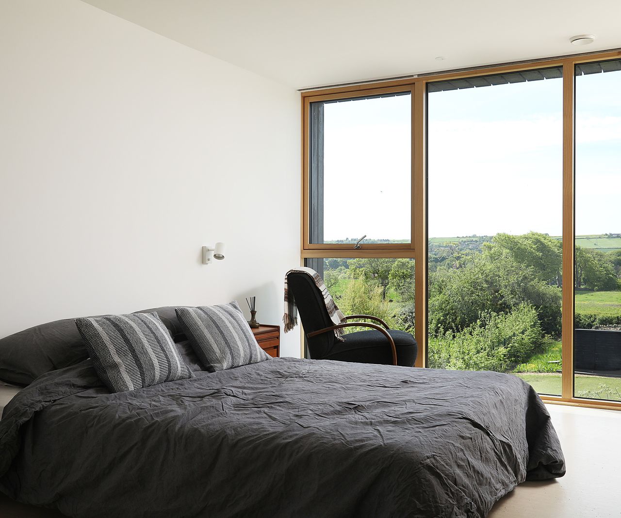 A double bed with crumpled cotton duvet cover in a dark grey colour. There is a wall of floor-to-ceiling-glazing looking out towards views of the countryside. A dark armchair sits next to the window angled outwards so the view might be enjoyed.