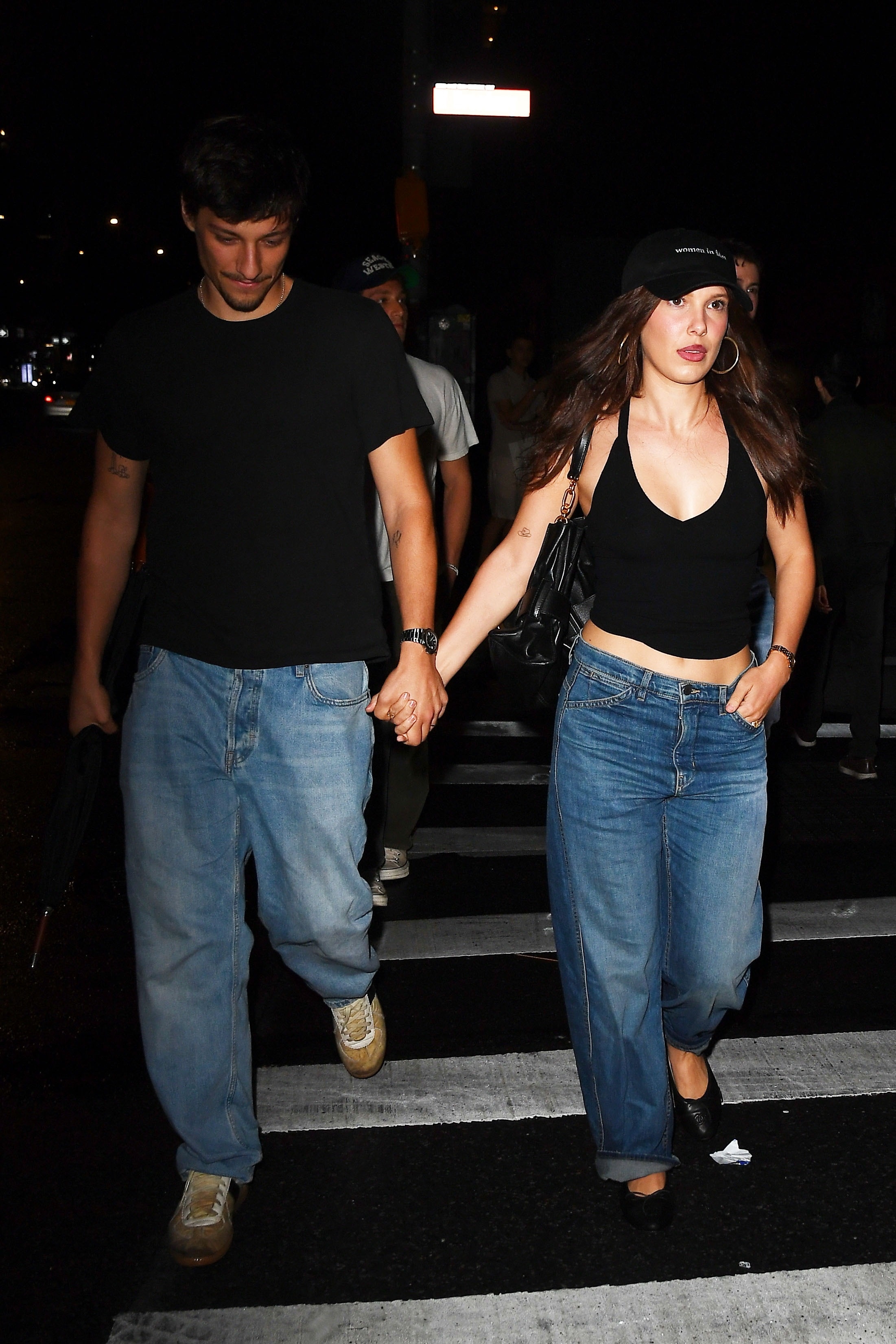 Millie Bobbie Brown walks down the street wearing cuffed jeans with a black halterneck.
