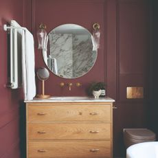 A burgundy-painted bathroom with wall panelling and a round hanging mirror above the sink