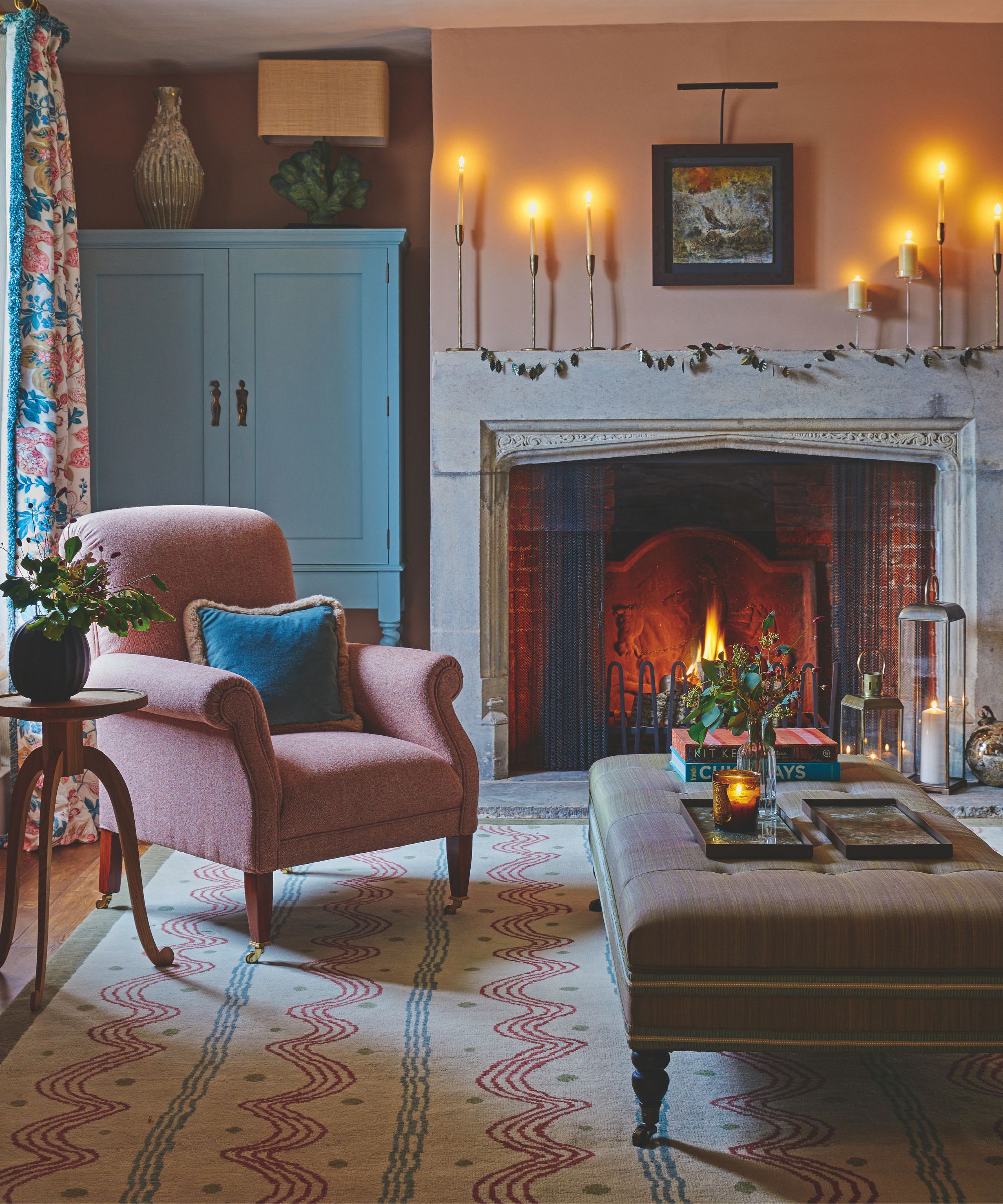 A pink and blue cozy living room with a lit fire in the large stone fireplace, candles, a patterned rug, pink chair, wooden sidetable, blue cabinet and large ottoman with a candle and vase of flowers.