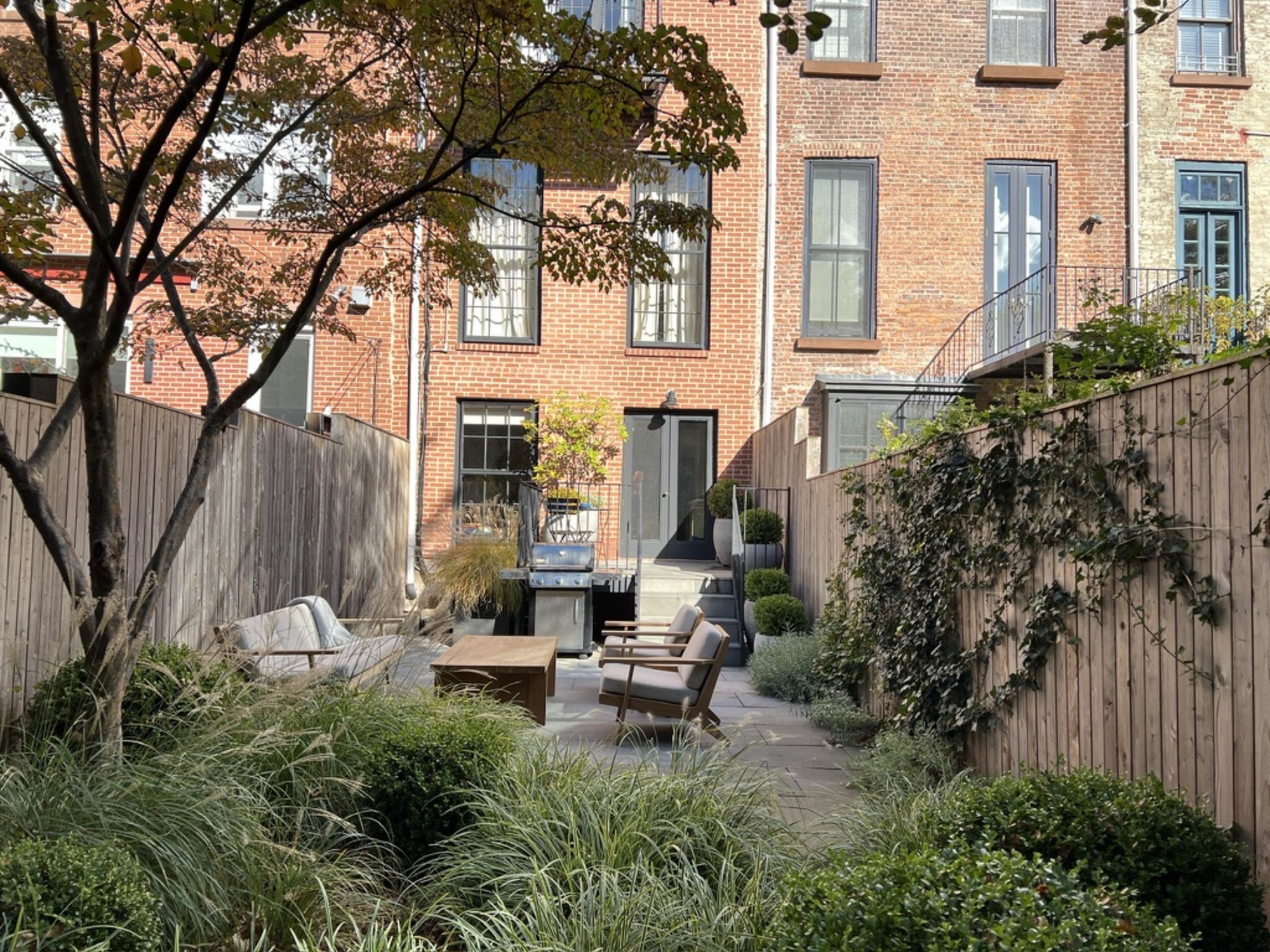 A garden of an urban terraced house with grasses in a flower bed and a garden furniture in the background