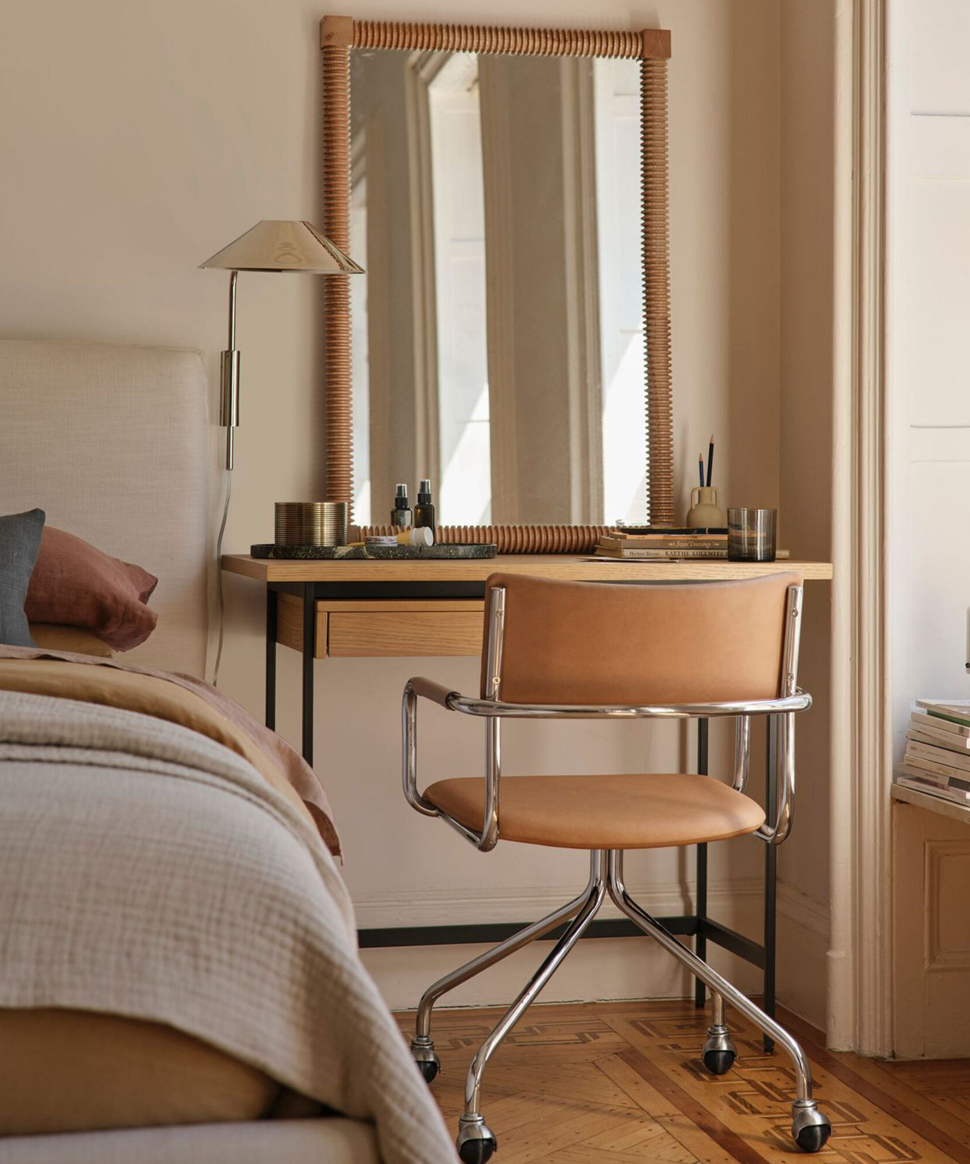 a bedroom with a small desk next to a bed, a desk chair, a mirror, and a plug in wall sconce on the wall