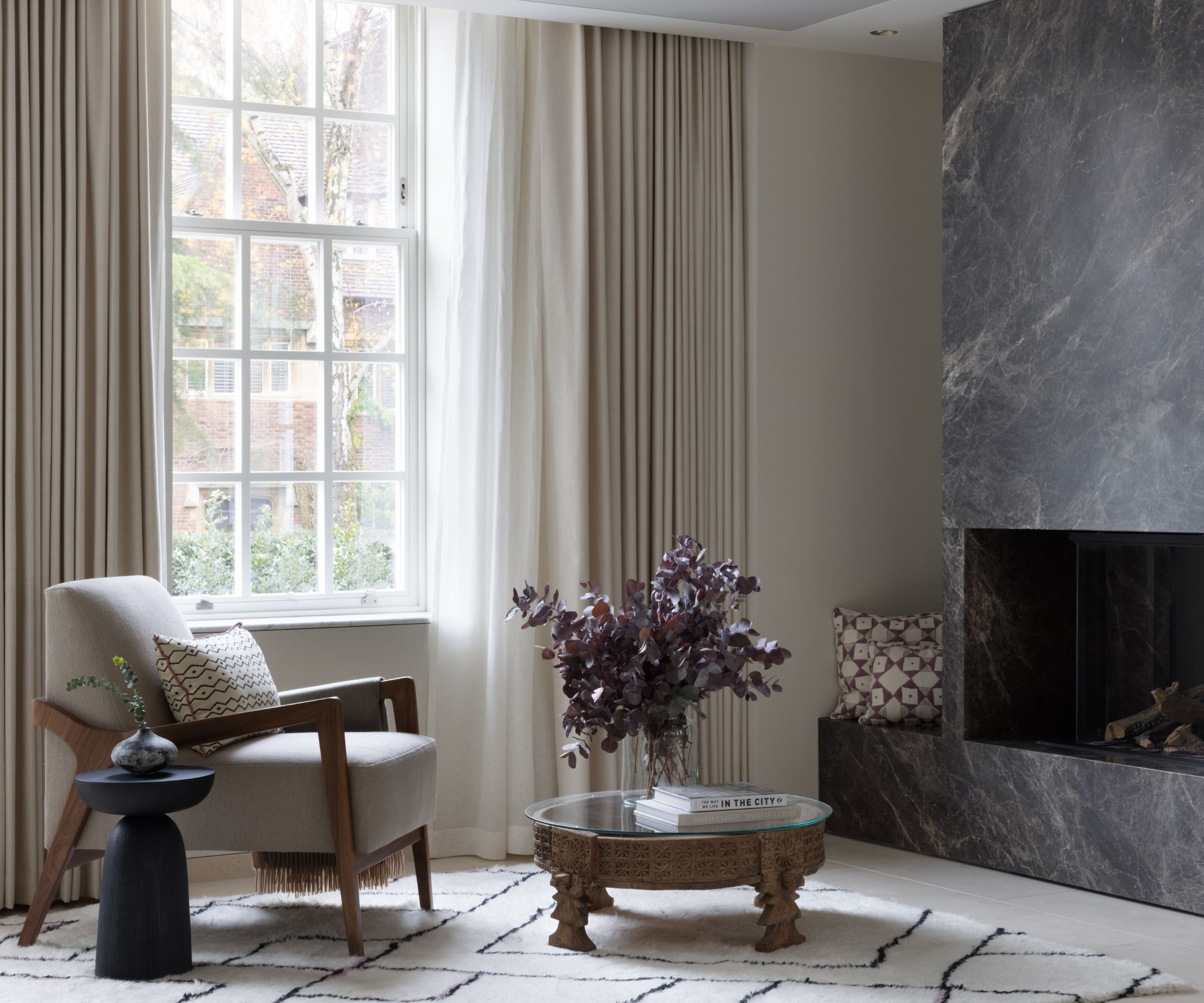 neutral living room with a white fluffy geometric rug, a dark grey floor to ceiling large marble fireplace, an antique wooden coffee table and a reading chair