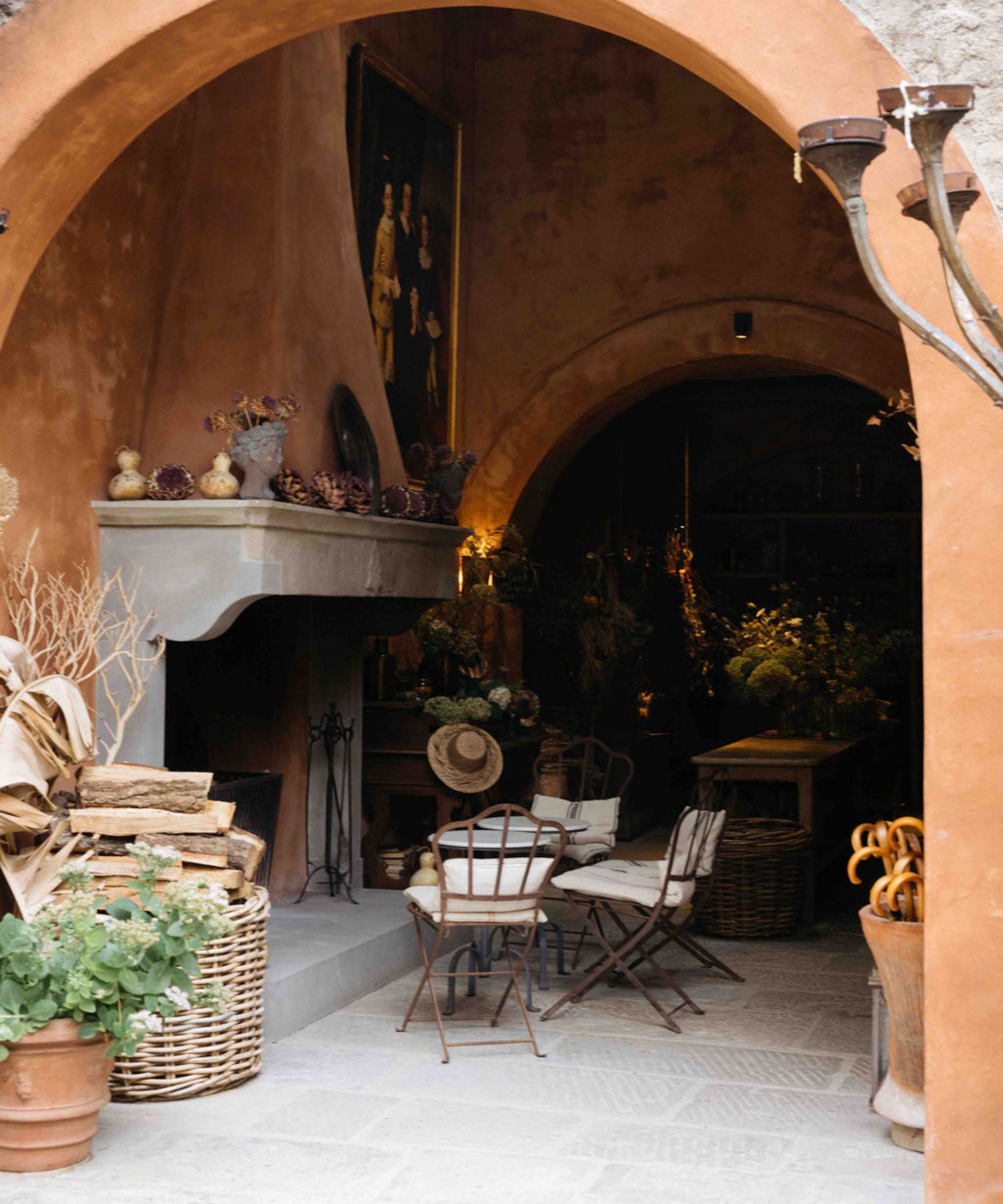 Image of inside an Italian shop, large stone fireplace, terracotta walls, seating and greenery, baskets of logs