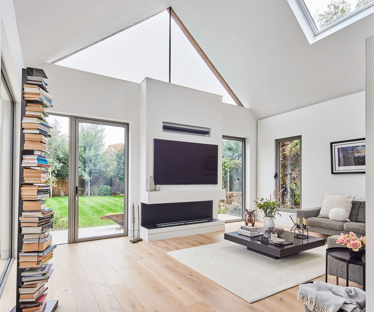 a living room with pitched ceiling and lits of architectural glazing. The floor is a light wood and the walls are white. There is a flat screen tv mounted on the wall above a contemporary fireplace